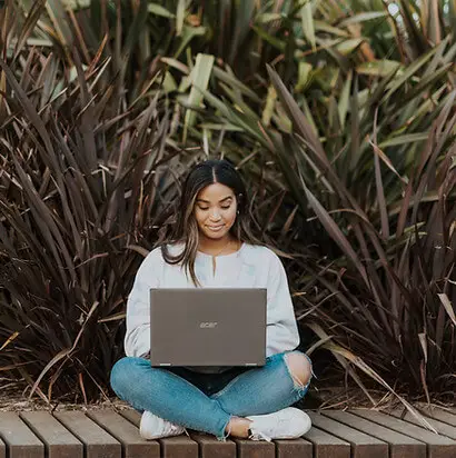 A woman sits cross-legged on a wooden deck, using her laptop to schedule mental health appointments. Surrounded by tall, leafy plants and dressed in a light jacket, jeans, and sneakers, her expression is focused yet relaxed.